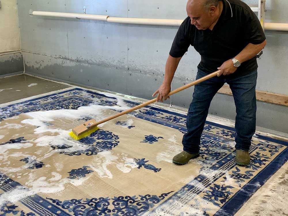 Un uomo strofina un tappeto blu e beige insaponato con una spazzola a manico lungo in una lavanderia che espone Lavaggio Professionale Tappeti a Melzo.
