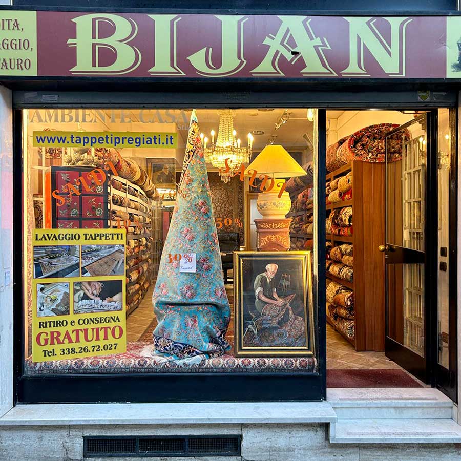 Vetrina con esposizione di tappeti arrotolati di Bijan Tappeti pregiati, una grande lampada, cartelli di vendita e un'opera d'arte raffigurante una donna che tesse.