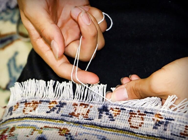 riparazione-tappeti-melzo-3 Primo piano di mani che intrecciano fili bianchi attraverso le frange di un tessuto fantasia, evidenziando il tocco artigianale che caratterizza i tappeti pregiati e la qualità associata a Bijan servizi.