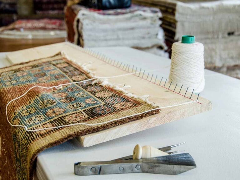 riparazione-tappeti-melzo Un tappeto parzialmente tessuto su un telaio con filo, una bobina e strumenti per tessere su un tavolo, che mette in mostra la maestria dietro Bijan Tappeti pregiati.
