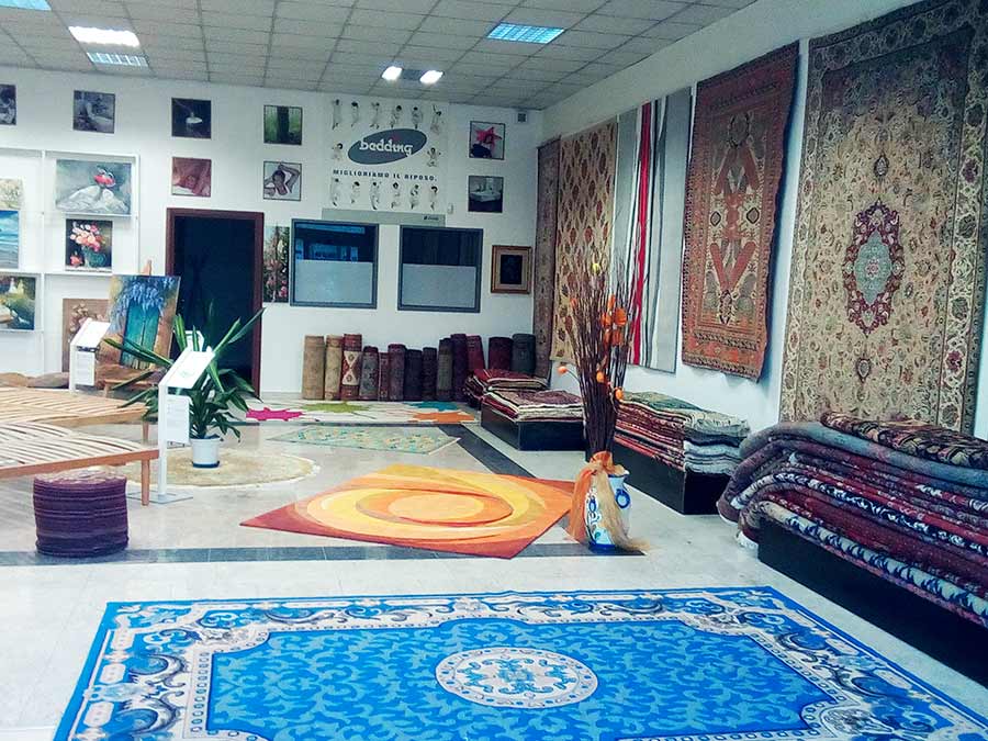 Showroom a Melzo che espone vari tappeti pregiati e colorati alle pareti e sui pavimenti, alcuni arrotolati e impilati, specializzato nella vendita tappeti.